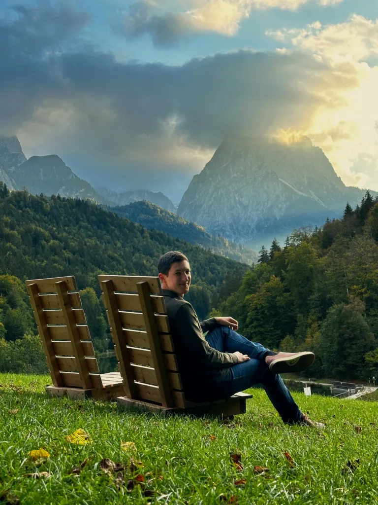 Hayden sitting in a wooden hillside chair overlooking an alpine lake and mountain peaks at sunset
